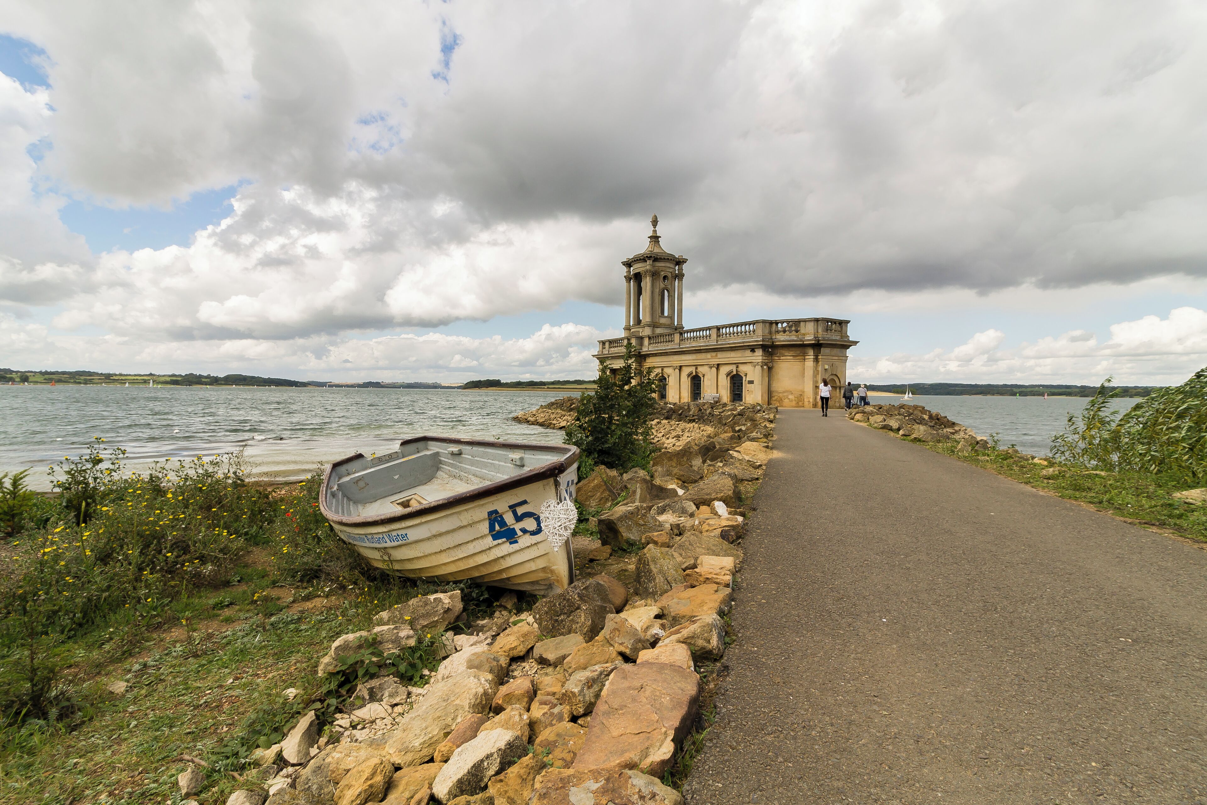 A coastal path with a rowboat, a stone wall, and a historic building with a tower.