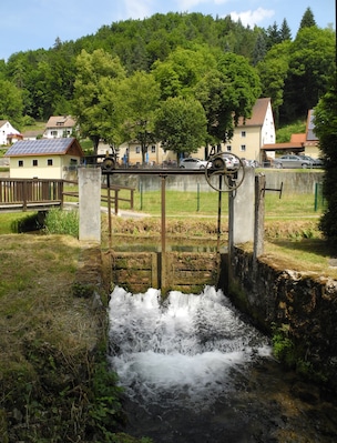 Uma pequena cascata em área rural, com casas e árvores ao fundo.