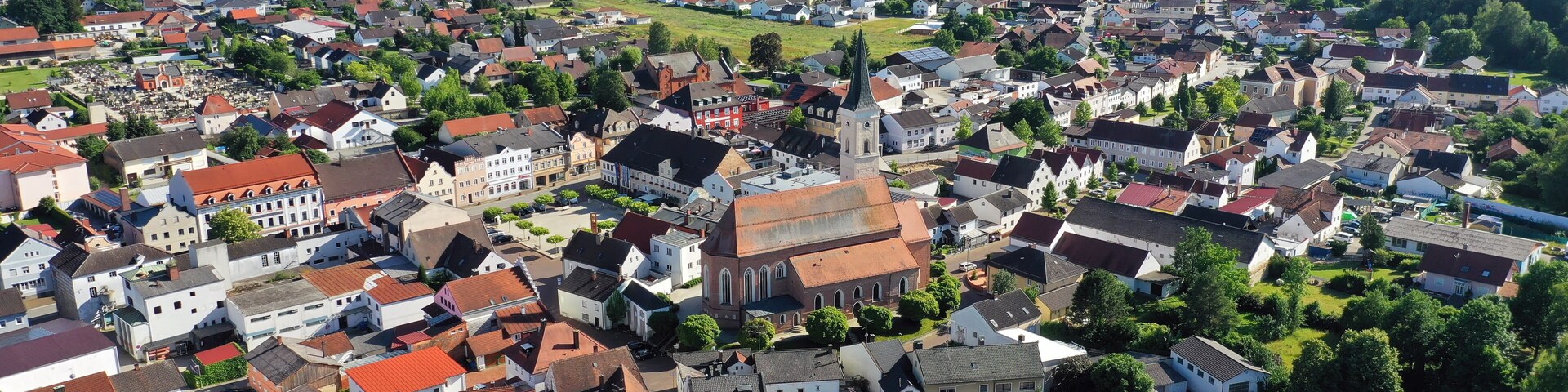 Vista aérea de uma cidade com uma igreja imponente, cercada por casas residenciais e campos verdes.