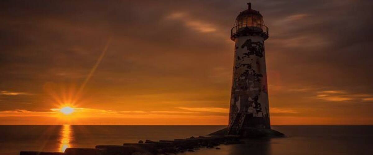 A lighthouse at sunset with the sun low on the horizon, reflecting on calm waters.