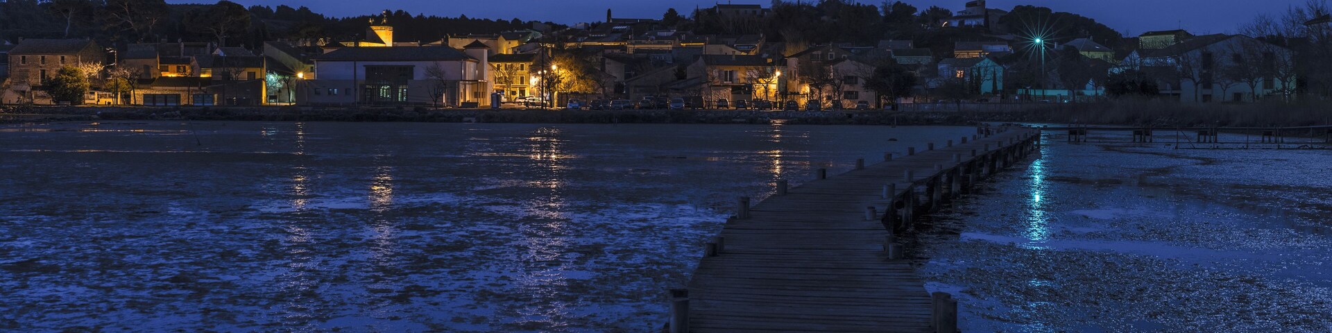 Um cais de madeira que se estende sobre a água ao entardecer, com edifícios iluminados ao fundo.