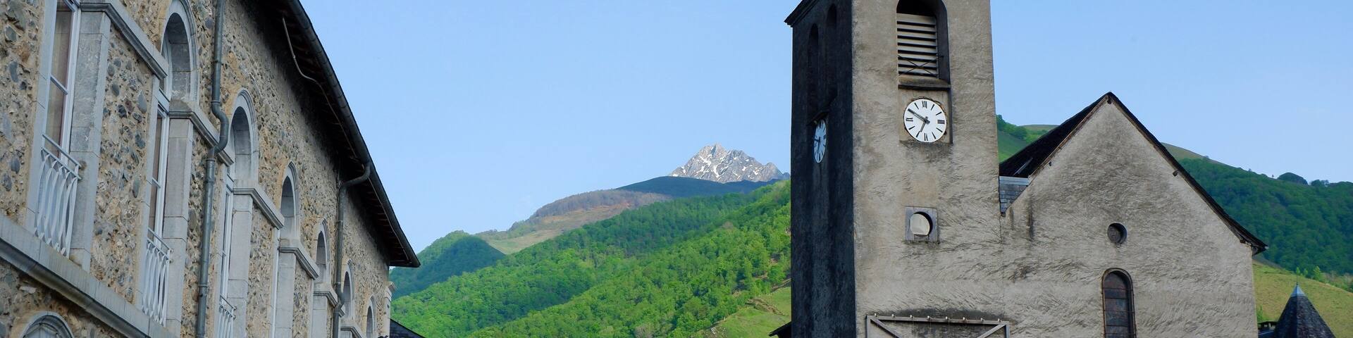 Charming square in a small village showcases a historic church with a clock tower alongside stone buildings in Bedous