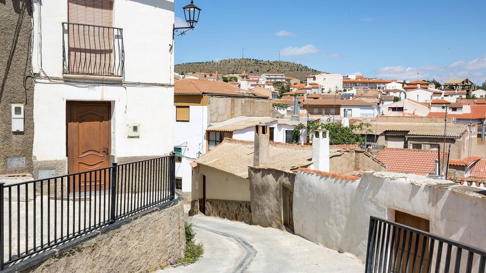 A narrow street in a Mediterranean town with terracotta-roofed buildings, a wooden door, and a metal railing.