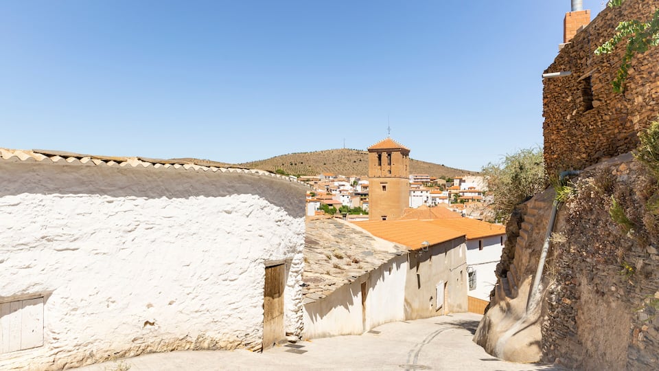 A narrow street with white and brown buildings, a tower, and a hill in the background.