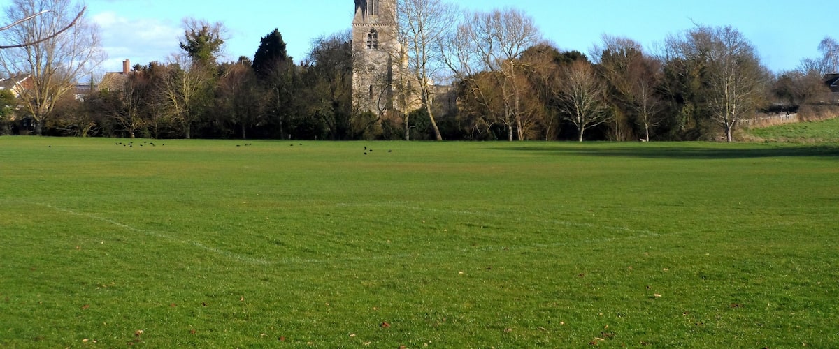 A church tower in a green field with trees and houses in the background.