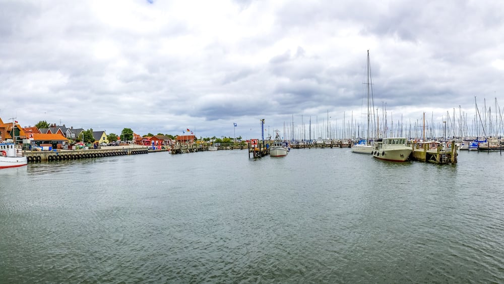 Ein Hafen mit zahlreichen Segelbooten, einem hölzernen Steg und Gebäuden im Hintergrund.