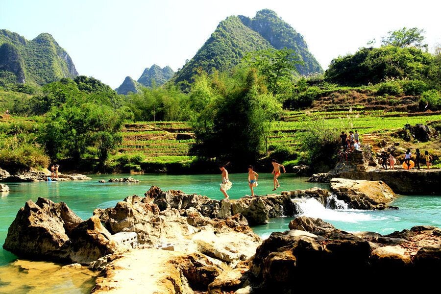 A river with rocky edges, people swimming and standing on rocks, and terraced fields in a mountainous landscape.