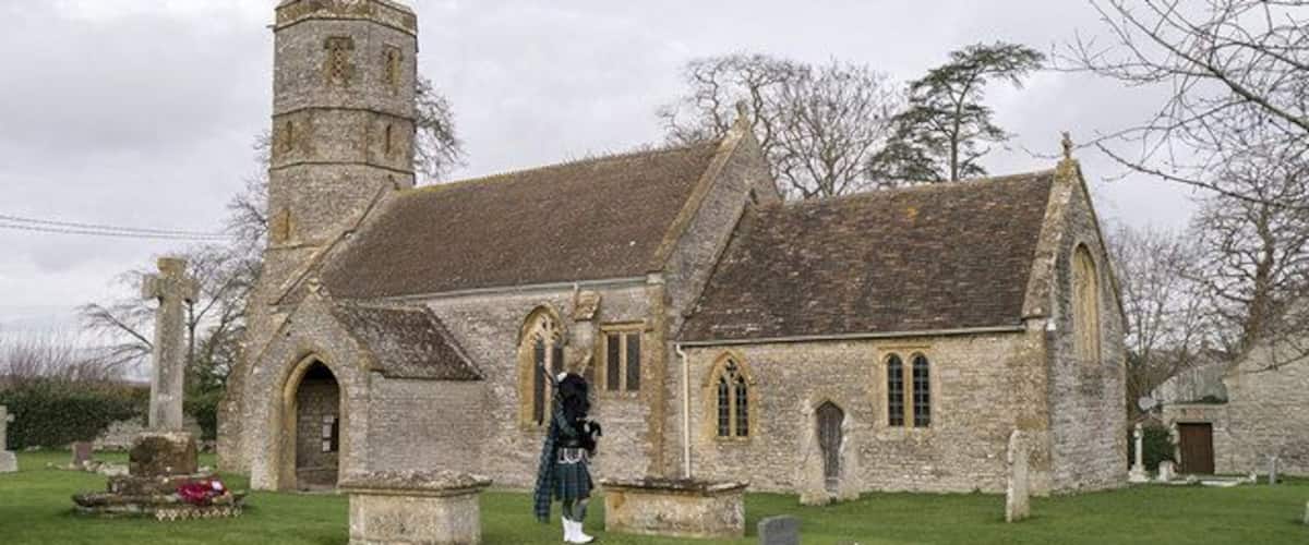 A stone church with a bell tower, a grassy cemetery, and a person walking.