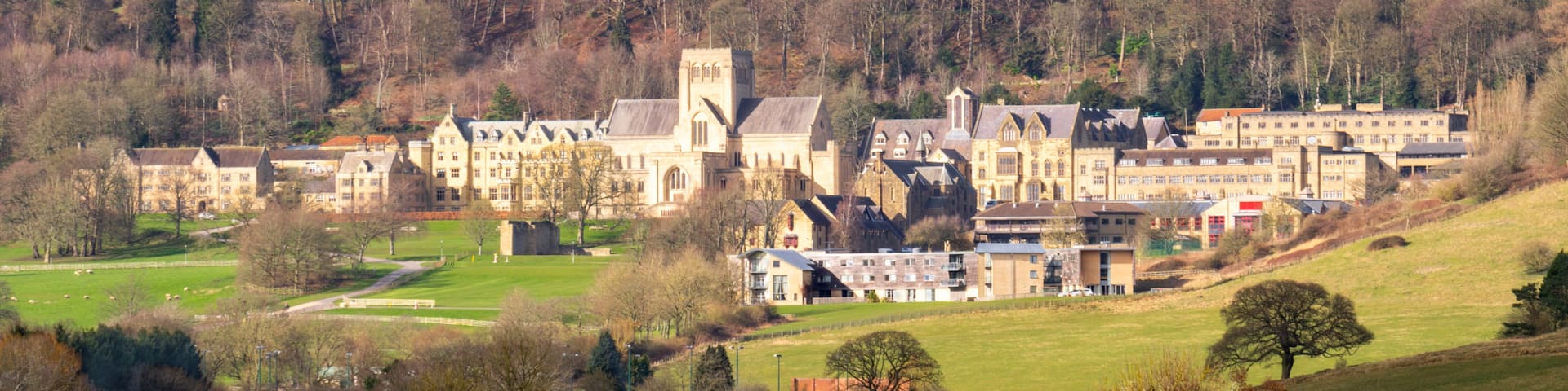 A historic building complex with a prominent tower, surrounded by rolling hills and a mix of deciduous trees.