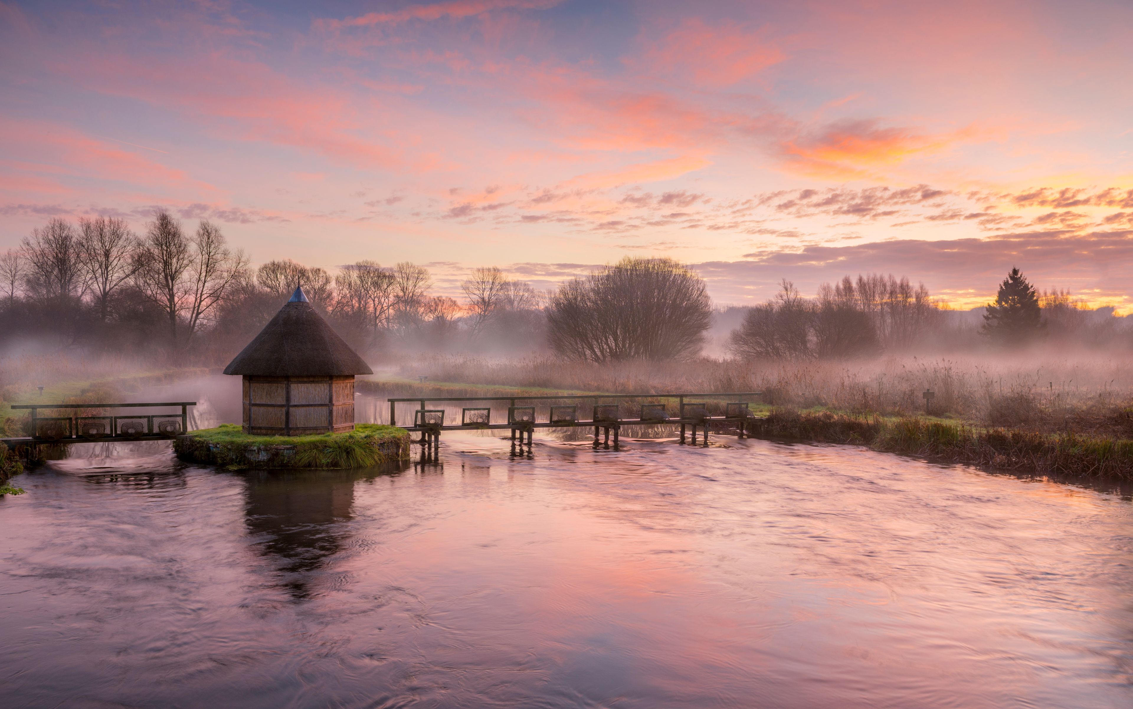 A thatched-roof hut by a calm river with mist, trees, and a sunset sky.