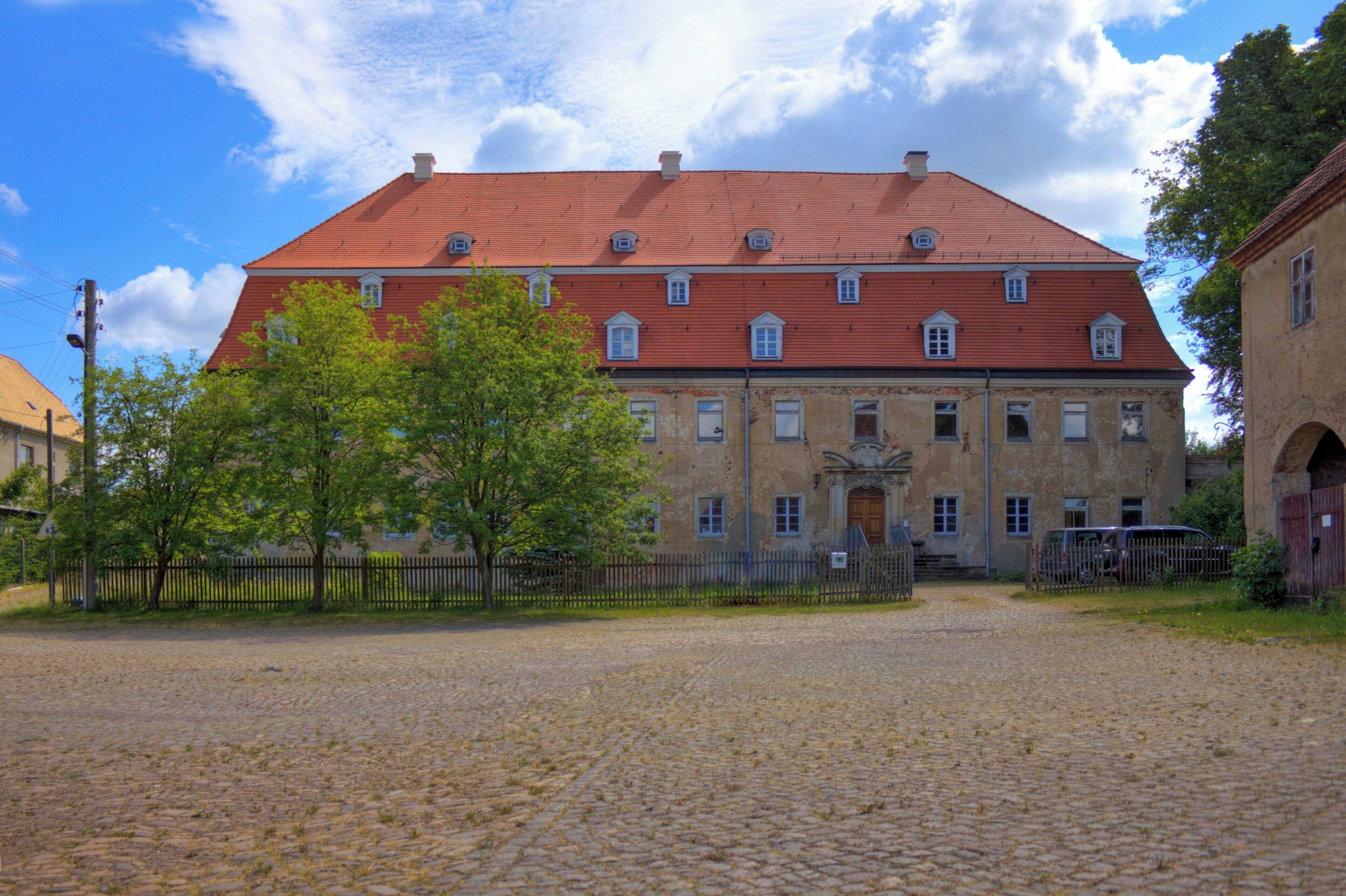A historic building with a red roof and multiple windows, surrounded by a cobblestone courtyard and a wooden fence.