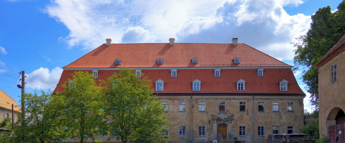 A historic building with a red roof and multiple windows, surrounded by a cobblestone courtyard and a wooden fence.