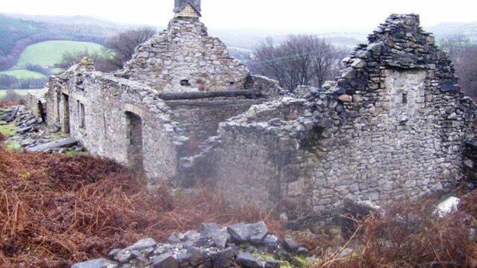 A ruined stone structure with a chimney, surrounded by a rural landscape.