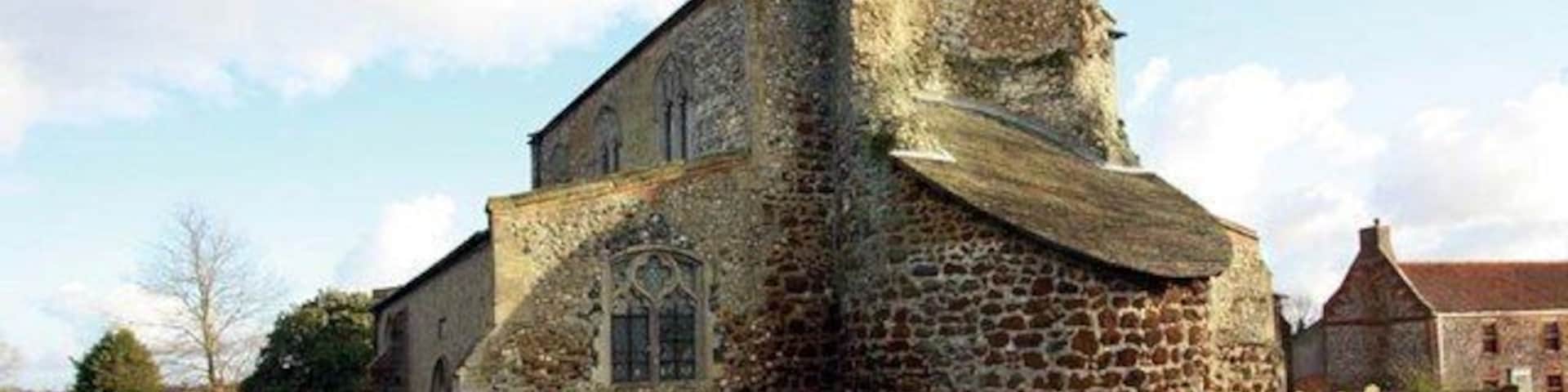 Una antigua iglesia de piedra con un techo empinado y un campanario, rodeada de un césped y otros edificios.
