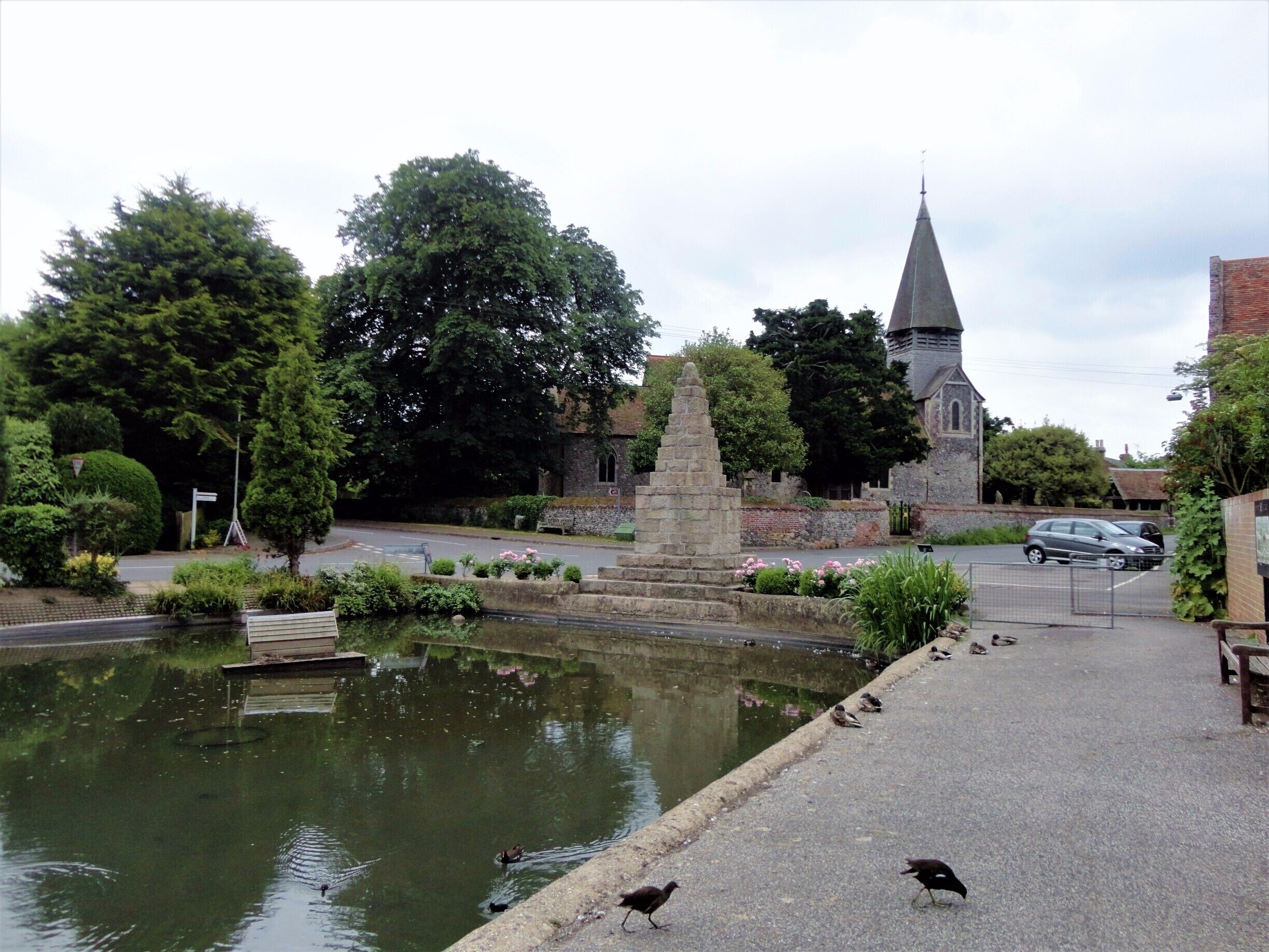 A pond with ducks, a stone monument, and a church with a steeple.