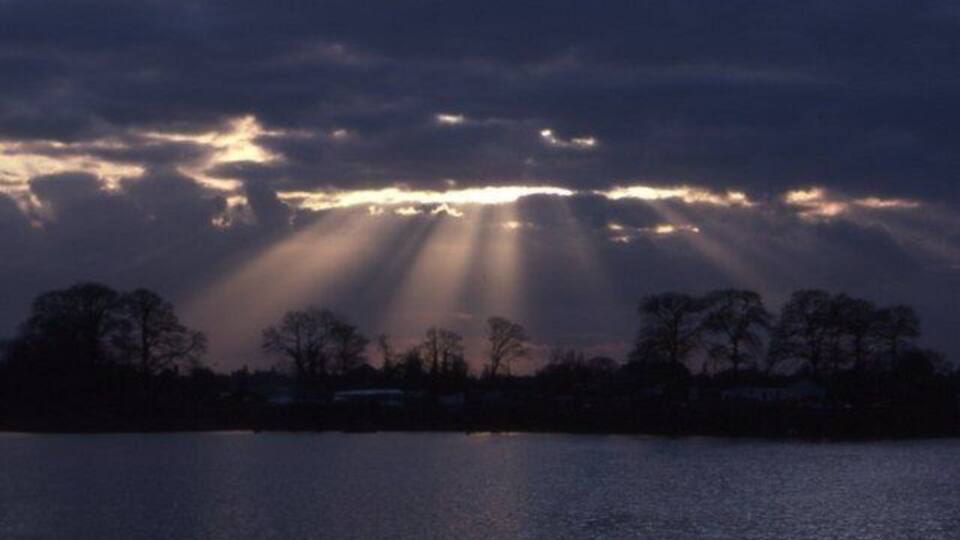 A lake with trees silhouetted against a cloudy sky, with sunlight streaming through the clouds.
