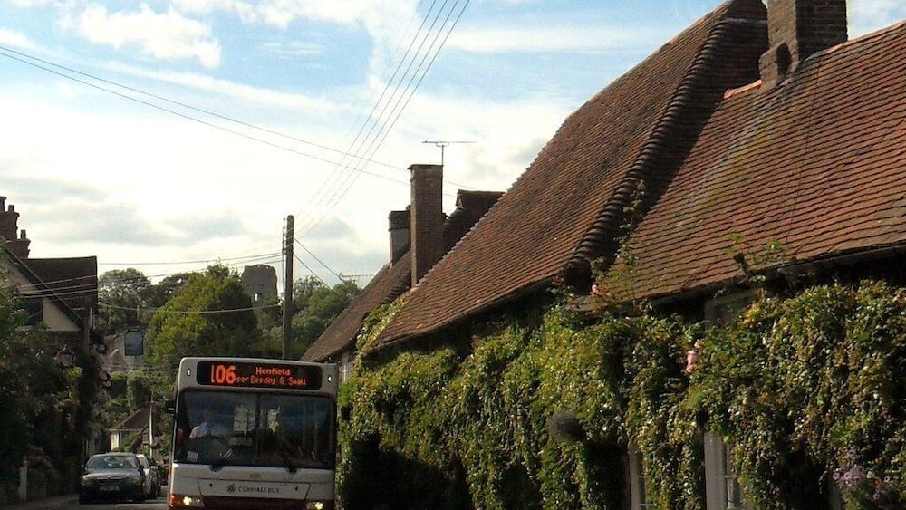 A bus on a rural road with houses covered in ivy and red flowers.