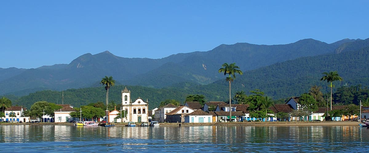 A coastal town with a church, boats, and palm trees against a backdrop of mountains.