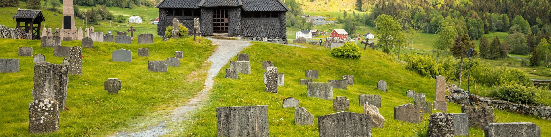 A church with a steeply pitched roof and a weather vane atop, surrounded by a graveyard with numerous headstones, set against a backdrop of a forested hillside under a cloudy sky.