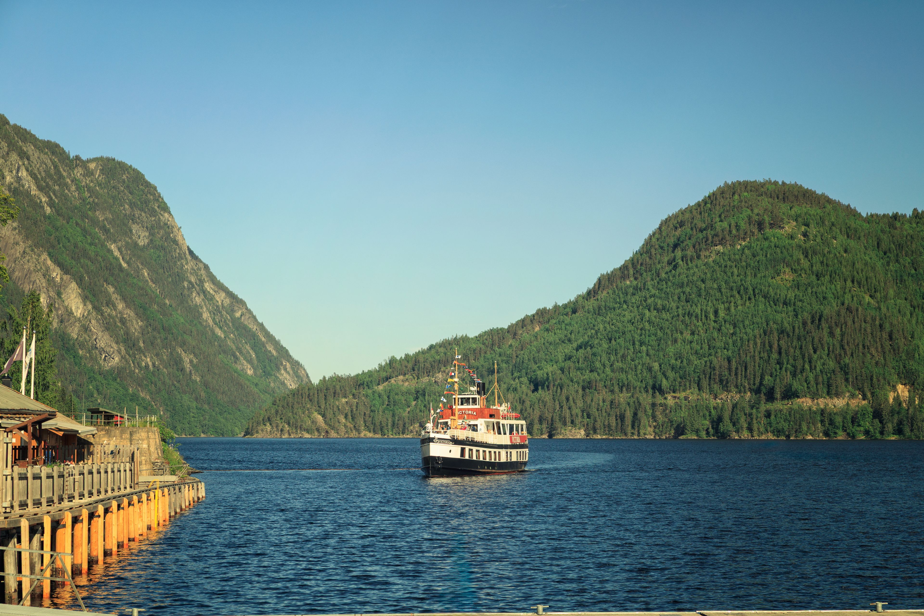 A boat named 'Victoria' on a calm lake surrounded by forested mountains.