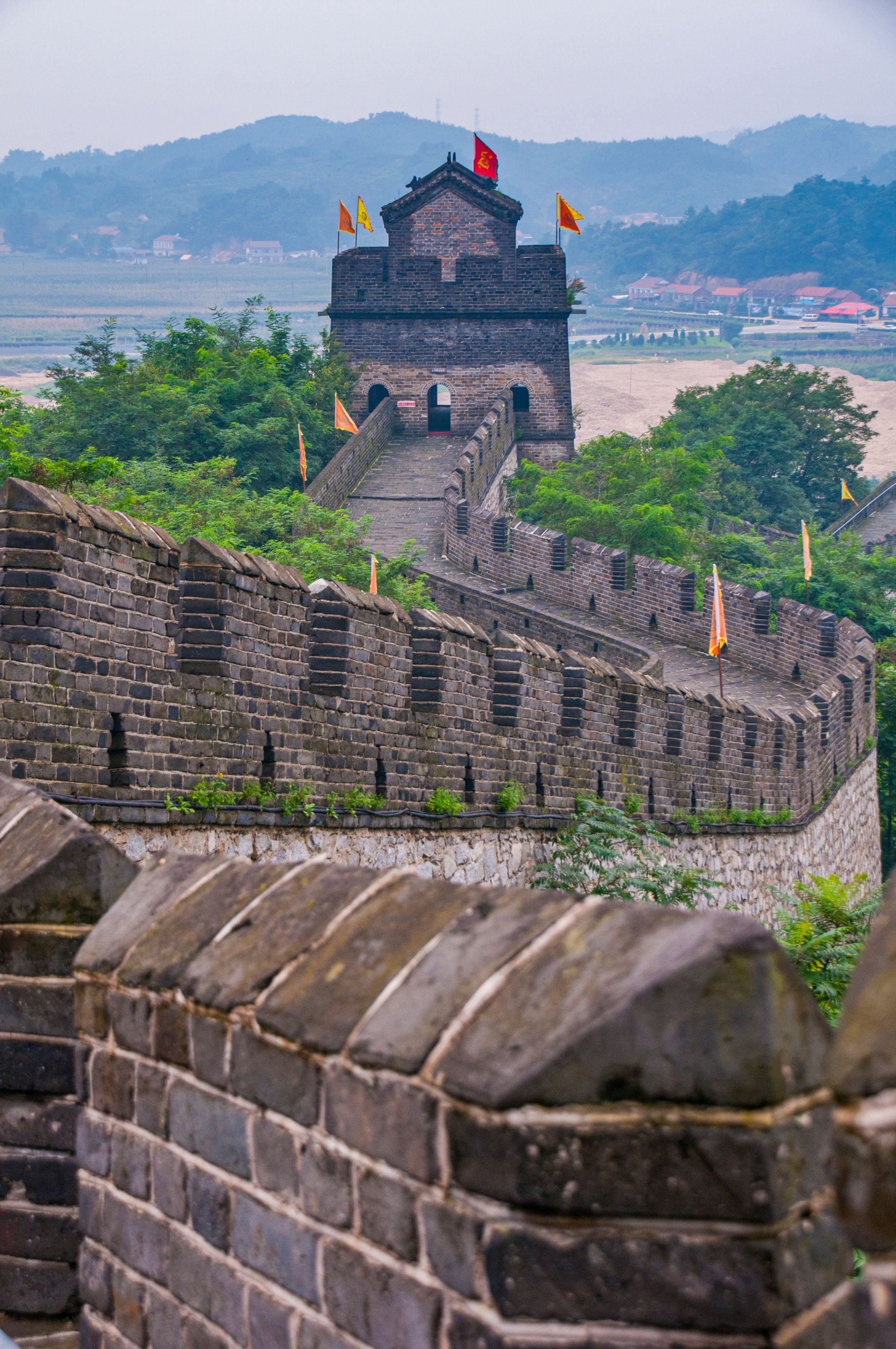 The Great Wall of China with watchtowers and flags, surrounded by lush greenery and distant mountains.