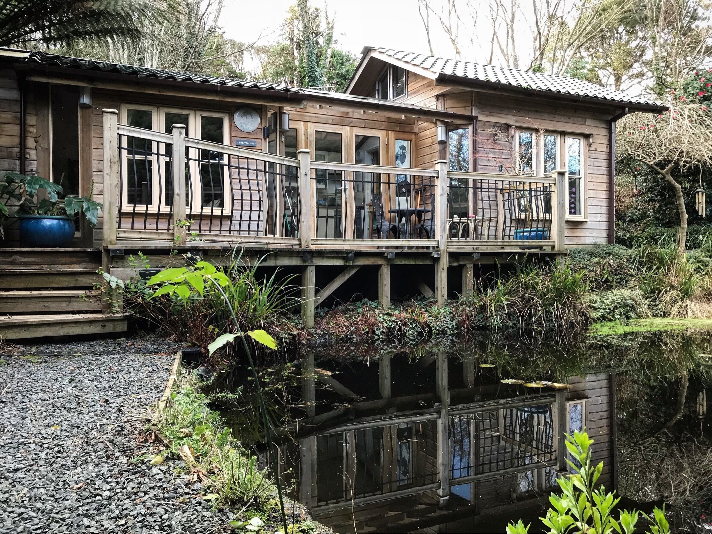 A wooden house with a balcony over a pond, surrounded by greenery.