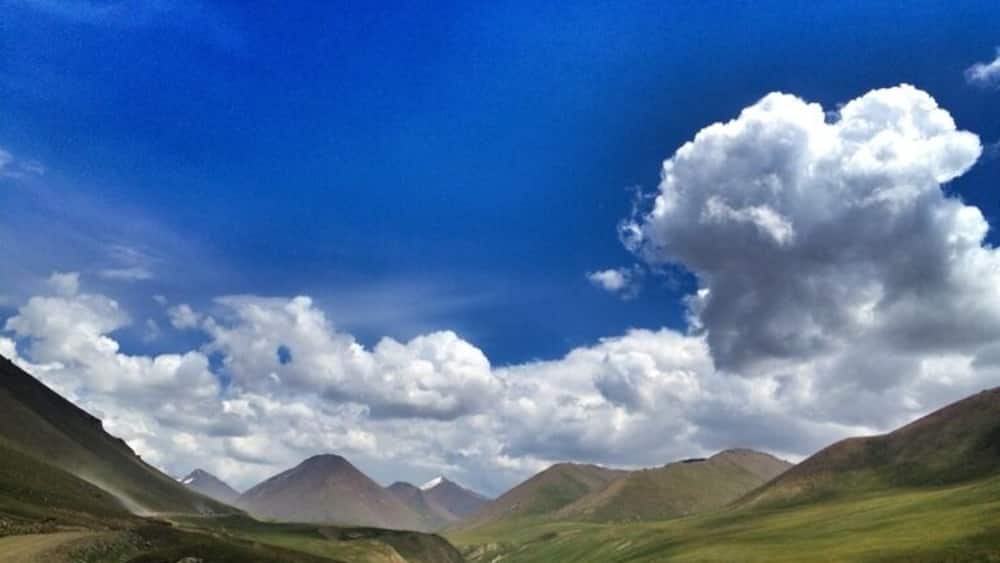 A mountainous landscape with a dirt path, rolling hills, and a large cloud in the sky.
