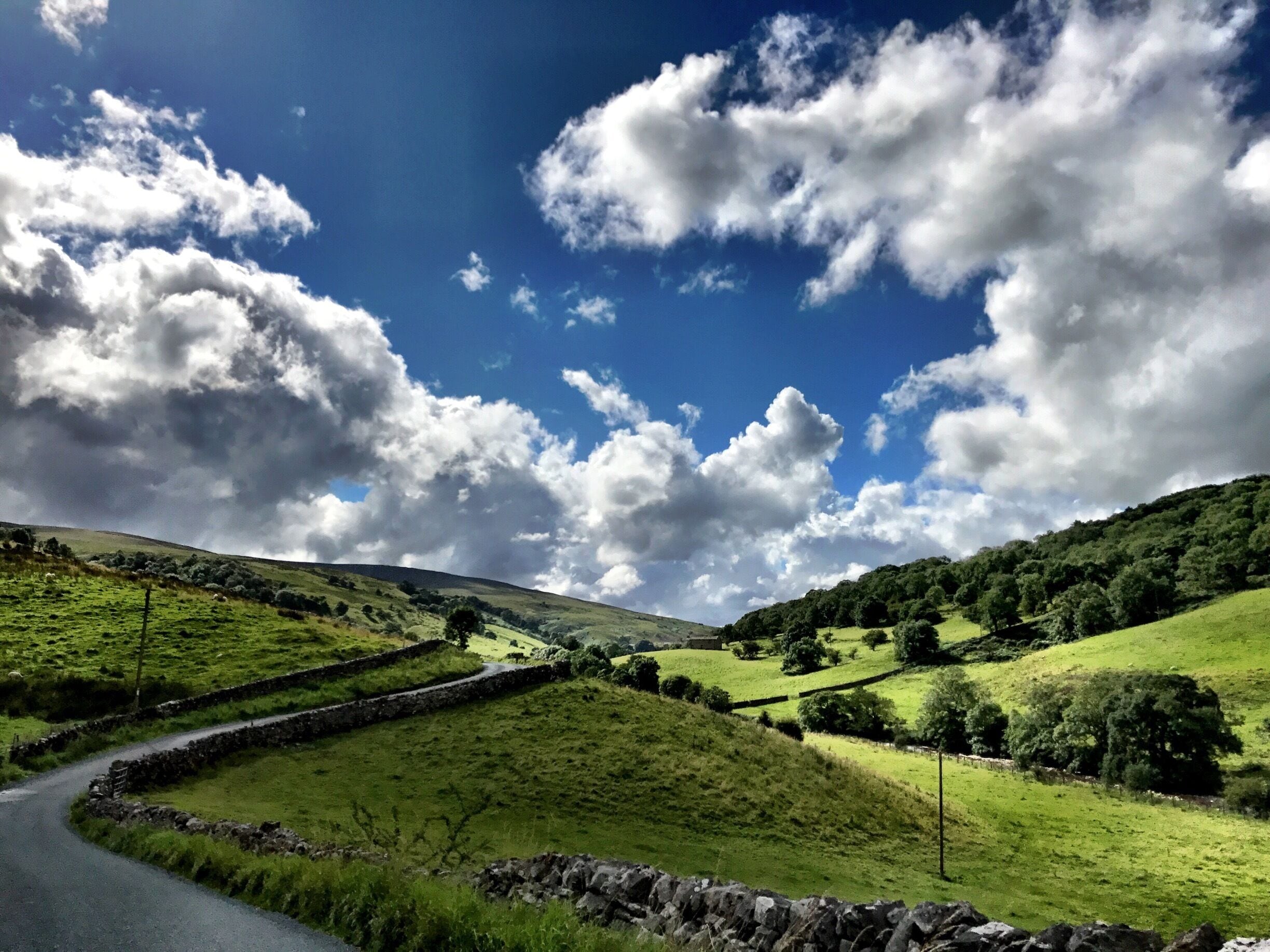 A winding country road through rolling green hills with stone walls and a clear blue sky with clouds.