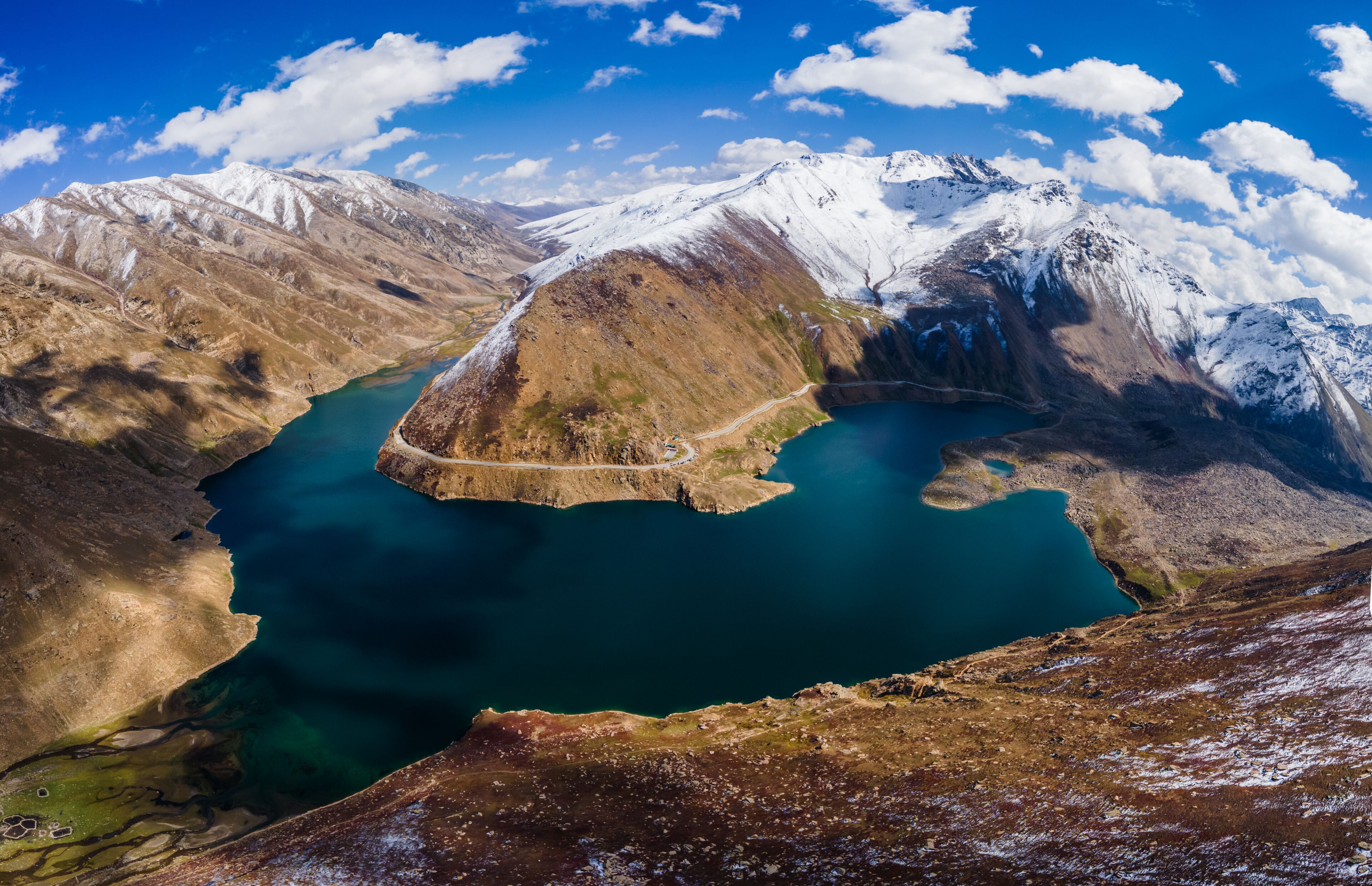 Aerial view of Lulusar Lake with deep blue waters surrounded by snow-capped mountain peaks and a winding road in Balakot, Khyber Pakhtunkhwa, Pakistan.
