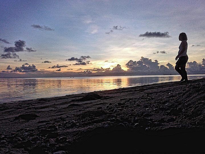 Una persona de pie en la playa al atardecer, con un mar tranquilo y un cielo nublado.