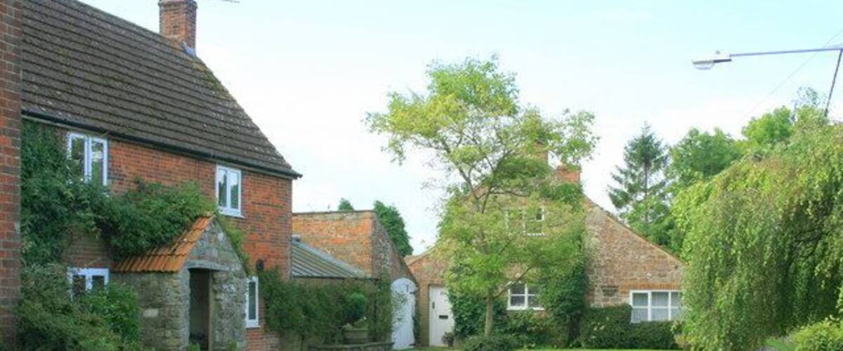 A quiet rural street with brick houses, a grassy area, and a tree-lined path.
