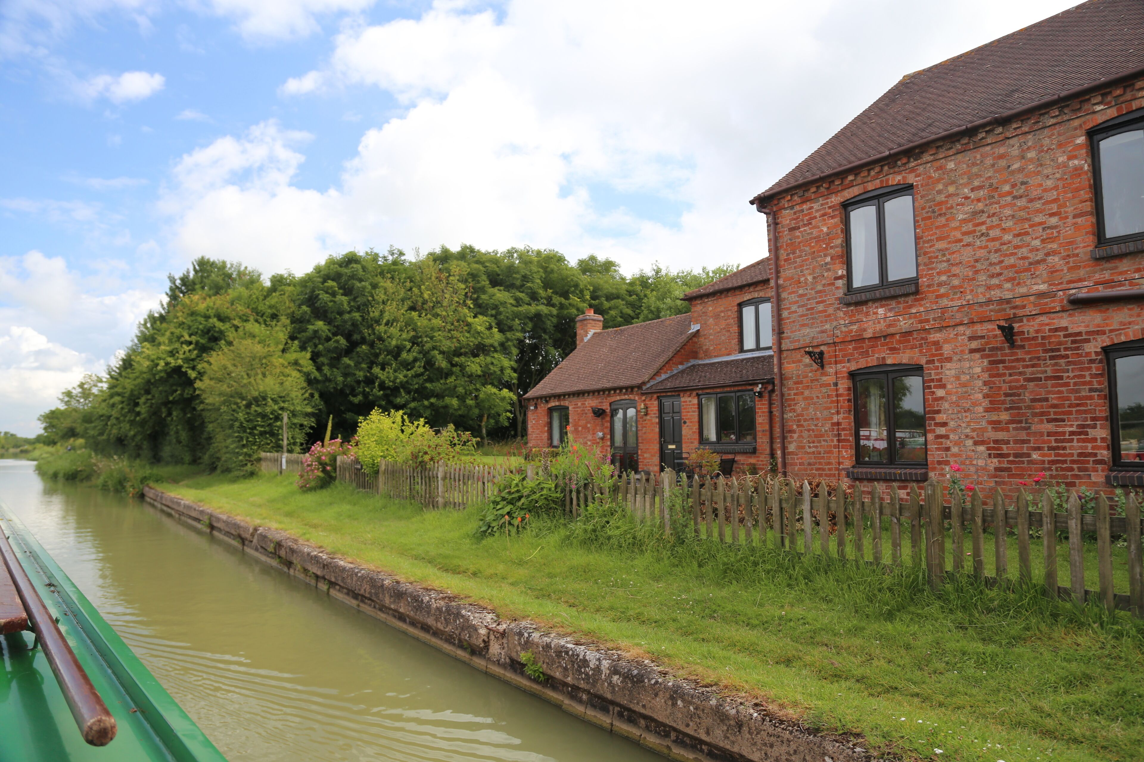 A canal with a green boat, a brick house, and a wooden fence.