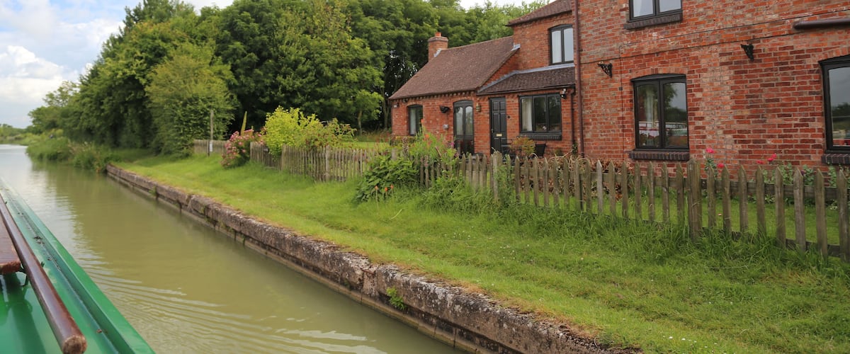 A canal with a green boat, a brick house, and a wooden fence.