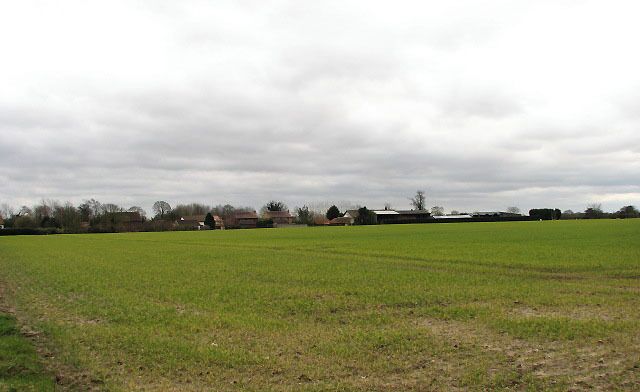 A rural landscape with a large open field, a few scattered buildings, and a cloudy sky.
