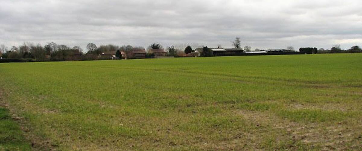 A rural landscape with a large open field, a few scattered buildings, and a cloudy sky.