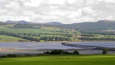 A coastal landscape with a bridge over water, green fields, and hills in the background.