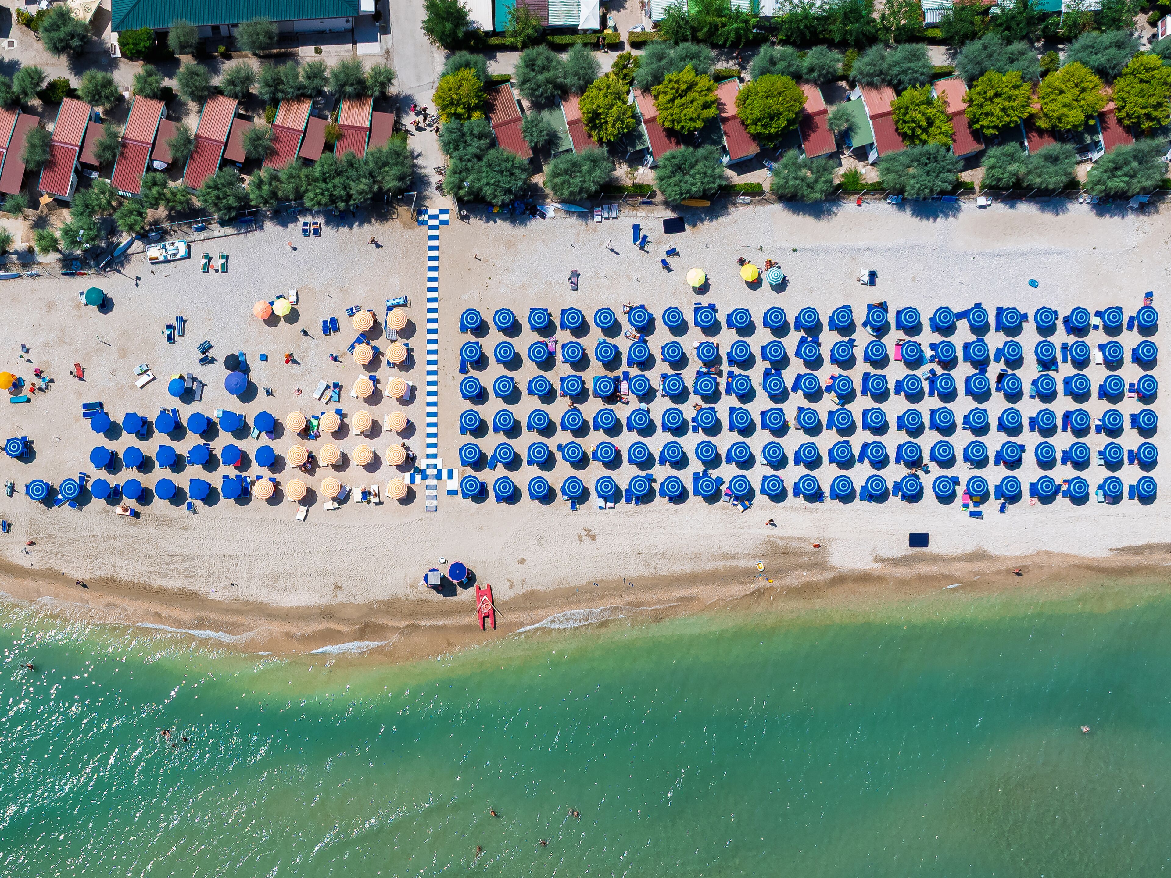 Aerial view of neat rows of blue umbrellas contrast the golden sand where the clear turquoise sea gently kisses the shore, Torre di Palme, Marche, Italy.