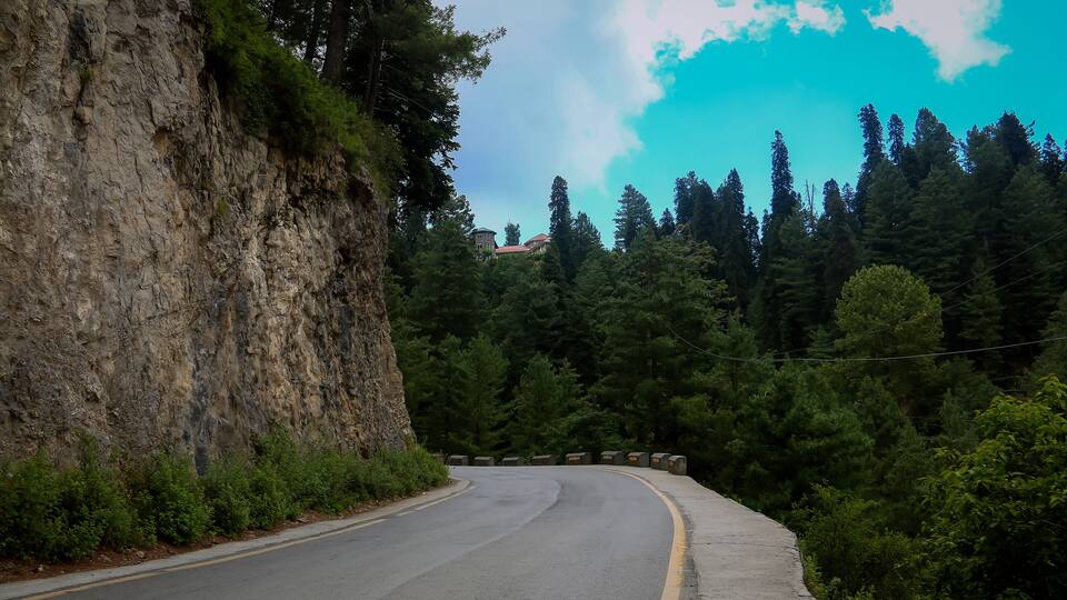 A winding road with a rocky cliff on the left and dense forest on the right.