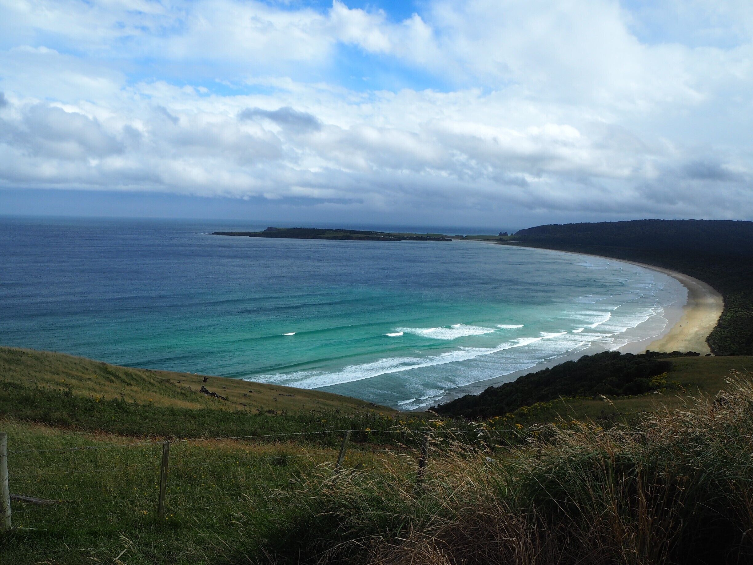 A coastal landscape with a sandy beach, ocean waves, grassy hills, and a distant island.