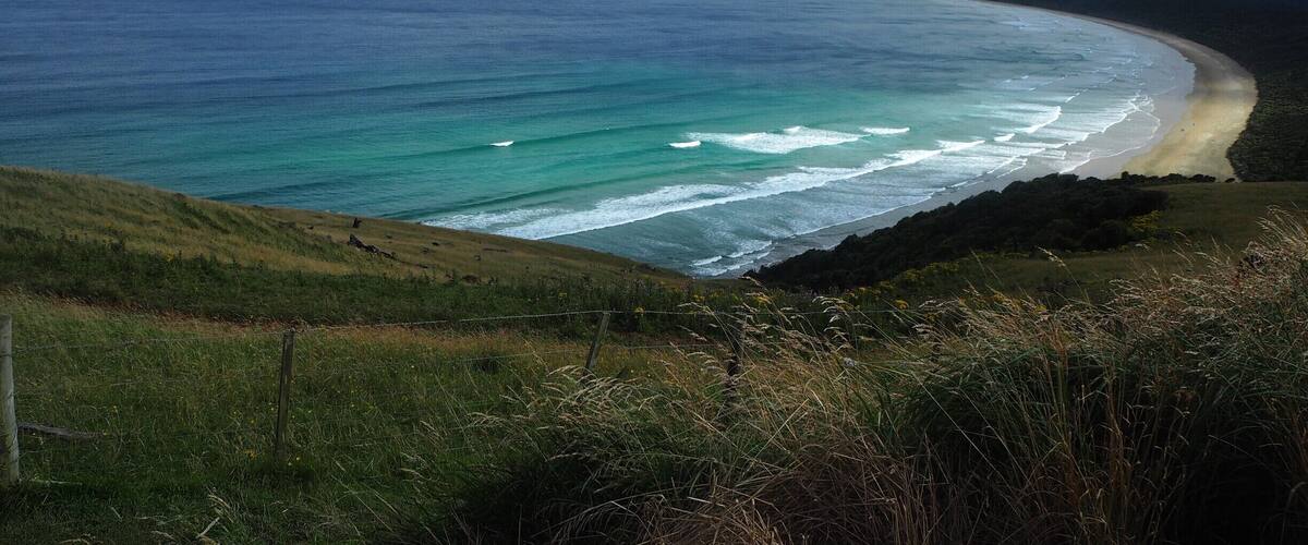 A coastal landscape with a sandy beach, ocean waves, grassy hills, and a distant island.