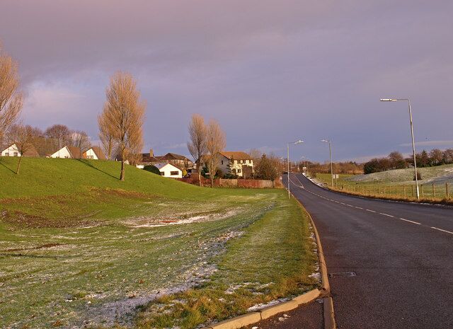 A rural road with houses on a hill, a grassy area, and a cloudy sky.