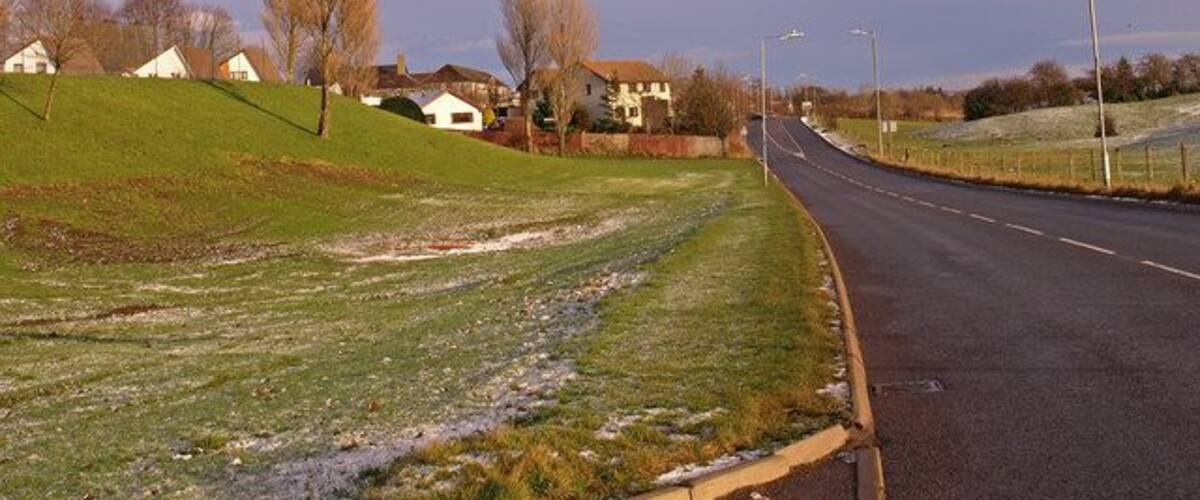A rural road with houses on a hill, a grassy area, and a cloudy sky.
