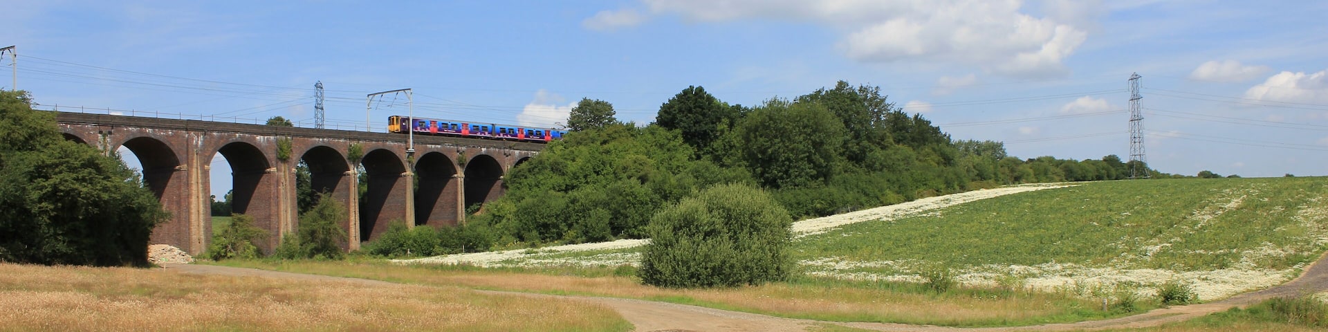 A train crossing a brick viaduct over a dirt road in a rural area with green fields and trees.