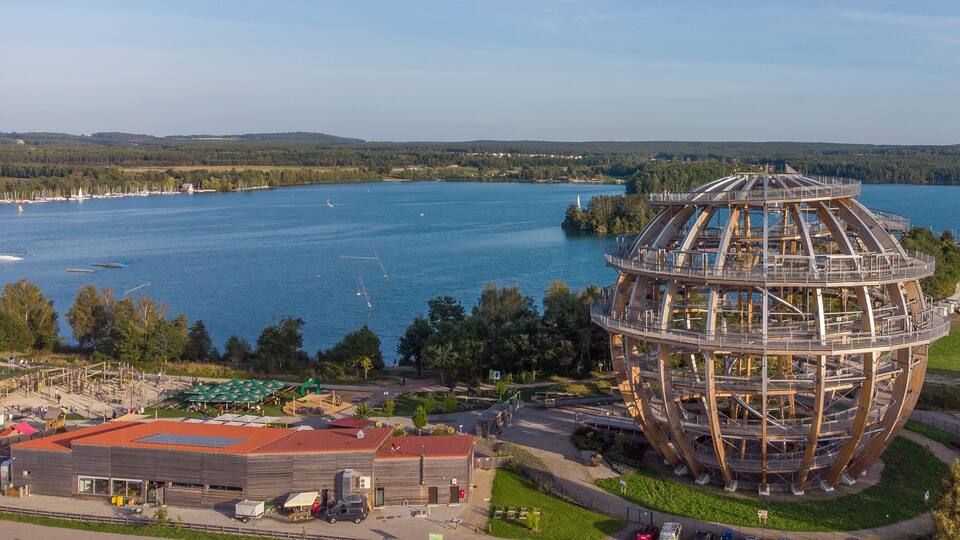 A large wooden observation tower by a lake with boats and a park nearby.