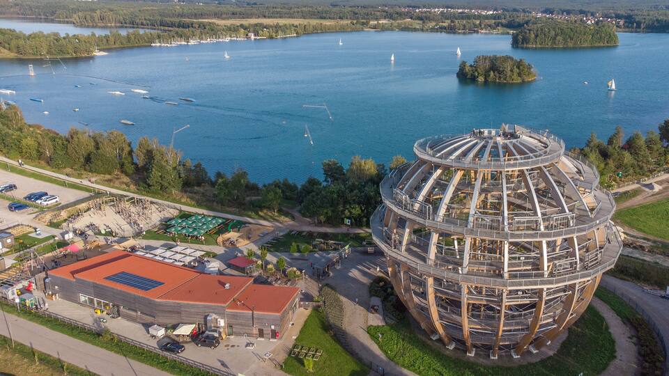 A large lake with sailboats, a circular building under construction, and a parking area with a red-roofed building.