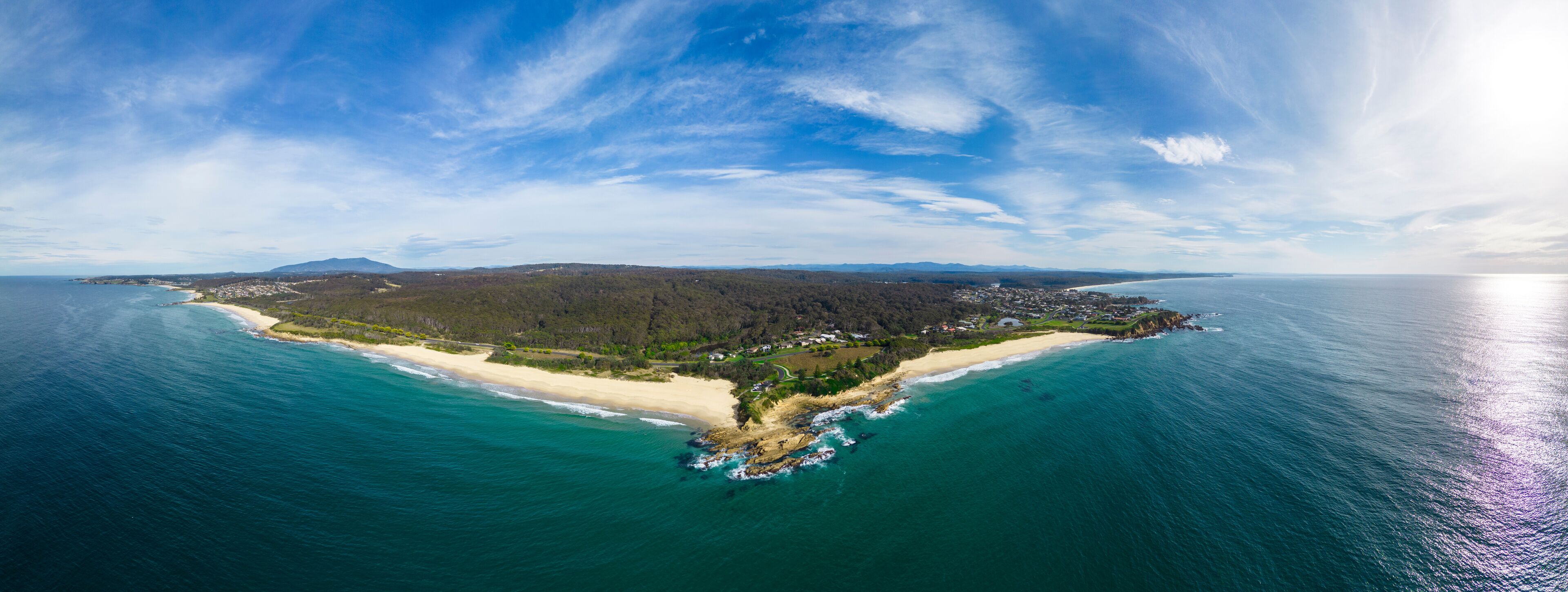 An aerial view of a coastal island with a sandy beach, dense forest, and a clear blue sky.