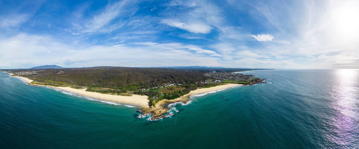 An aerial view of a coastal island with a sandy beach, dense forest, and a clear blue sky.
