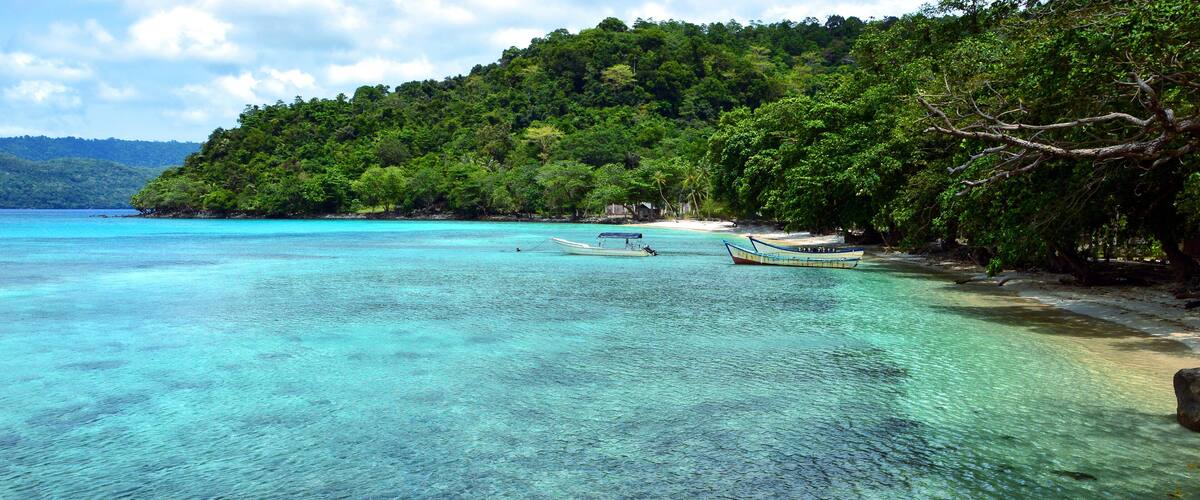 A clear turquoise sea, a sandy beach, and a lush green hill with boats in the water.