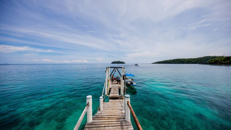 A wooden pier extending into turquoise waters, leading to a small island with a boat docked nearby.