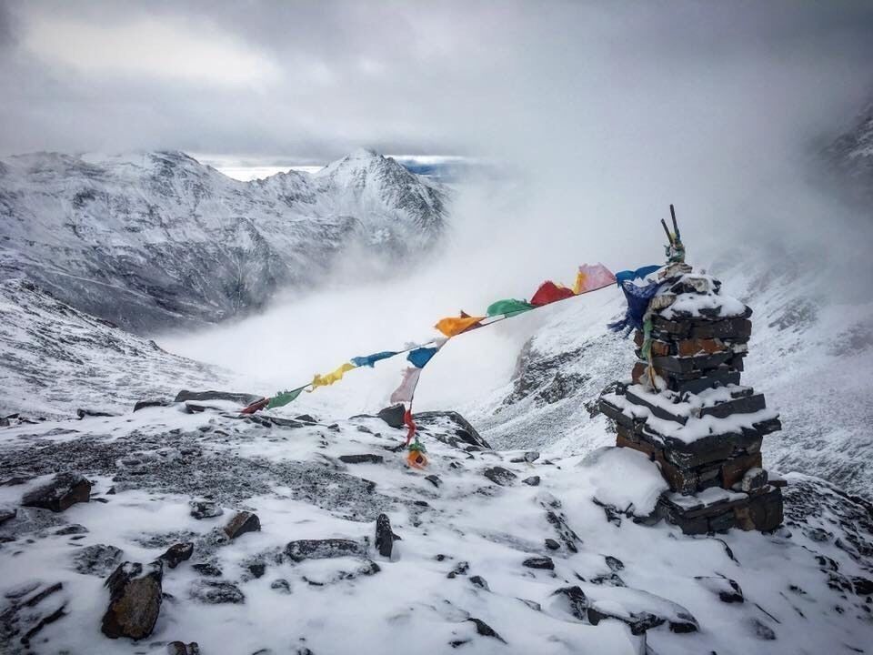 A snow-covered mountain peak with a small shrine and colorful prayer flags.